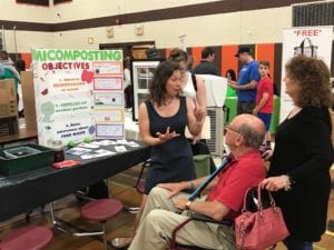 Three people discuss residential composting while looking at a student poster in a school gym.