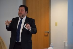 A man in a suit stands before a room of people and speaks during a presentation