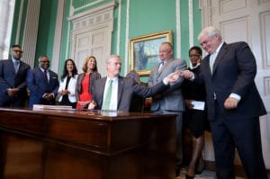 Mass Governor Charlie Backer sits at a desk with 7 people behind him, handing a pen to a man on the right