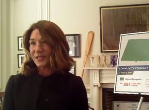 a woman speaking into a computer camera with a presentation board behind her.