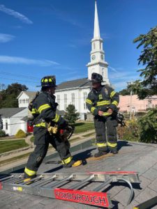 two firefighters stand on a roof with a ladder next to them, a church steeple in the background.