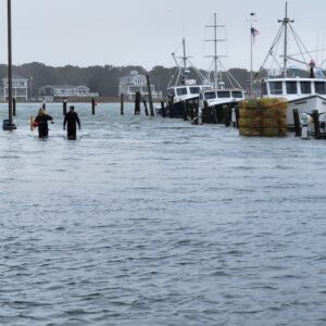 two people in wet suits walk in flood water next to ships docked in harbor
